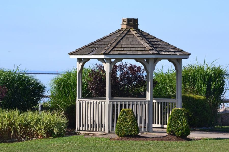 Timber garden gazebo among mature planting beside calm water in daylight