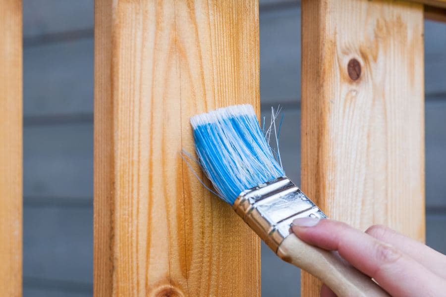 Hand applying clear sealer to a timber upright with a paintbrush against a gray wall